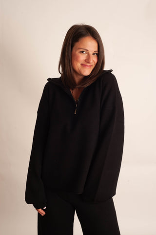 A woman with shoulder-length brown hair wearing a black half-zip pullover hoodie and black pants, standing against a plain off-white studio background.