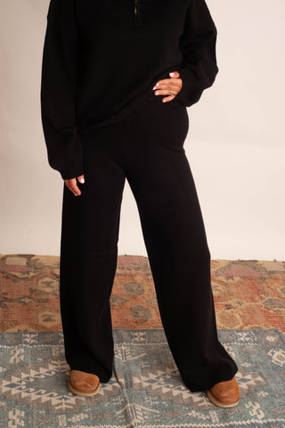 A person wearing a black lounge set consisting of a long-sleeve half-zip pullover and matching wide-leg pants, paired with brown suede shoes, standing on a patterned rug against a plain light background.