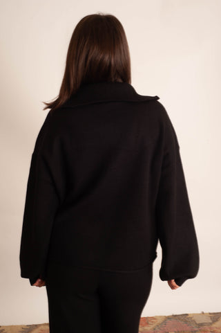 A person viewed from the back wearing a loose black long-sleeve top or jacket with a wide collar, paired with black pants, standing against a plain off-white wall.