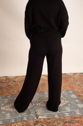 A person shown from the back wearing a black long-sleeve top and black wide-leg pants, standing on a patterned rug against a plain light wall.