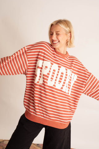 A woman wearing a rust-orange and white striped crewneck sweatshirt with 'FROCK' printed in large white letters across the chest, paired with black pants, posing against a plain off-white wall.