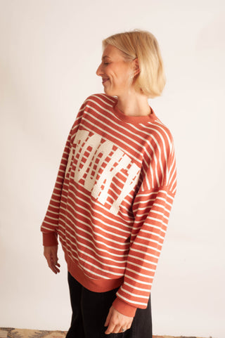 A woman with short blonde hair is shown from the side, wearing a rust-orange and white striped crewneck sweatshirt with a large white graphic on the front, paired with black pants, against a plain off-white wall.