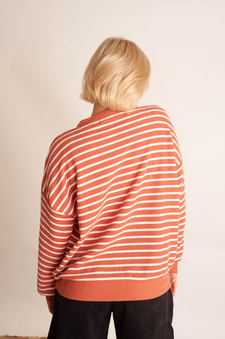 A person viewed from behind wearing a loose, long-sleeve knit sweater with thin horizontal orange and white stripes, paired with dark pants, against a plain off-white wall.