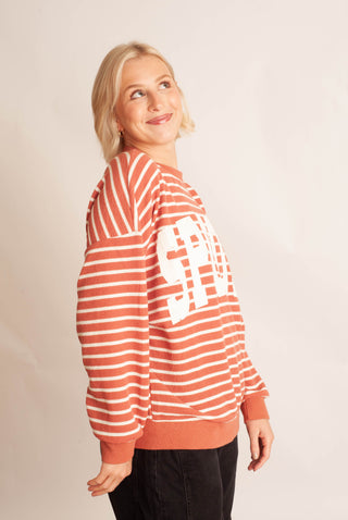 A woman wearing a rust-orange and white striped crewneck sweatshirt with a large white graphic on the front, paired with black pants, photographed against a plain light beige studio background.