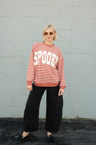 A woman stands against a light gray painted brick wall wearing a rust-orange and white horizontally striped sweatshirt with 'SPOOKY' printed in large white letters across the chest, paired with black wide-leg pants and black loafers.