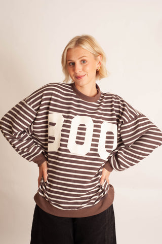 A woman with short blonde hair wearing a brown and white horizontally striped sweatshirt with large white '300' text across the chest, paired with black pants, posing against a light beige studio background.