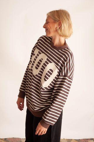 A woman with short blonde hair is shown from the side, wearing a brown and white striped long-sleeve sweater with a large cream-colored '100' graphic on the front, paired with black pants.