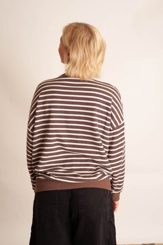 A person viewed from the back wearing a brown and off-white horizontally striped long-sleeve top and black pants, standing against a plain off-white wall.