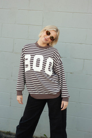 A woman wearing a brown and white horizontally striped long-sleeve sweatshirt with large white 'BOO' lettering on the front, paired with black pants, standing against a light gray painted brick wall.