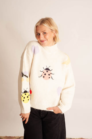 A woman modeling a cream-colored knit sweater with colorful embroidered ladybugs and abstract spots, paired with black pants, against a plain off-white background.