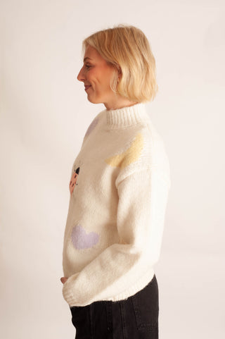 A woman with short blonde hair is shown in profile wearing a fuzzy cream-colored knit sweater with pastel yellow and purple graphic details, paired with black pants, against a plain off-white background.
