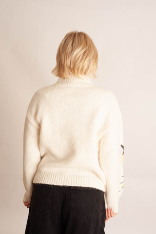 A person shown from the back wearing a cream-colored knit sweater and black pants, standing against a plain light beige studio background.