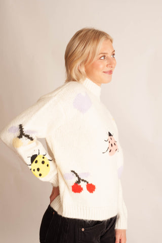 A woman wearing a cream-colored knit sweater with colorful cartoon ladybugs and cherries, paired with black pants, photographed against a plain light background.