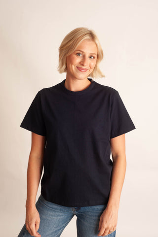 A woman with short blonde hair wearing a plain black short-sleeve crewneck t-shirt and blue jeans, standing against a light beige studio background.
