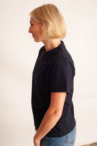 A woman with short blonde hair is shown in profile wearing a dark navy blue short-sleeve t-shirt and blue jeans against a plain off-white background.