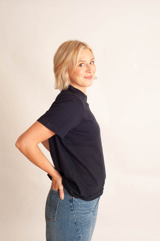 A woman with short blonde hair is shown from the side, wearing a dark navy blue short-sleeve polo shirt and blue jeans, standing against a plain off-white background.