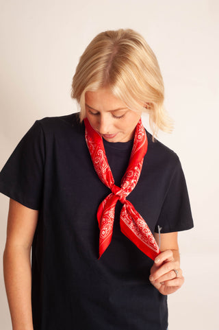 A blonde woman wearing a black t-shirt and a red bandana tied around her neck, looking down against a plain light beige background.