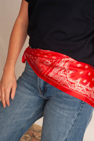 A close-up of a person wearing a bright red bandana with a white paisley pattern tied around the waist over blue jeans and a black top.