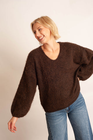 A smiling blonde woman wearing a fuzzy, oversized dark brown V-neck sweater and blue jeans, posing against a plain light beige studio background.