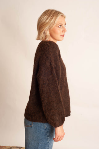 A woman with blonde hair is shown from a side-back angle wearing a fuzzy, textured dark brown knit sweater and blue jeans against a plain off-white background.