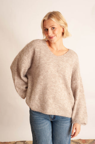 A woman with blonde hair wearing a fuzzy, oversized light beige knit sweater and blue jeans, standing against a plain off-white background.