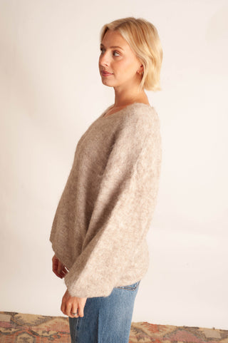 A woman with short blonde hair wearing a fuzzy, oversized beige knit sweater and blue jeans, standing in profile against a plain off-white wall.