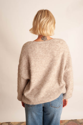 A woman with short blonde hair is shown from behind wearing a fuzzy, oversized light beige knit sweater and blue jeans against a plain off-white wall.
