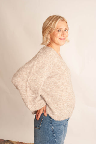A woman with blonde hair wearing a chunky, oversized knit sweater in a heathered light beige color, paired with blue jeans, posing against a plain off-white studio background.