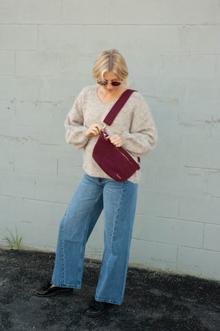 A woman wearing a beige knit sweater, blue wide-leg jeans, and black flats carries a burgundy quilted crossbody bag against a light gray painted brick wall.