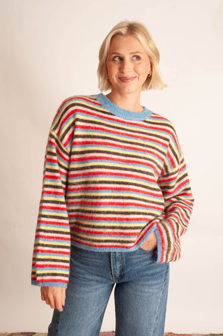 A woman with short blonde hair wearing a fuzzy, multicolored horizontally striped sweater and blue jeans, standing against a plain off-white background.