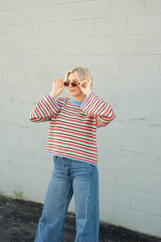 A woman wearing a colorful horizontally striped knit sweater with blue jeans and sunglasses, standing against a white painted brick wall.
