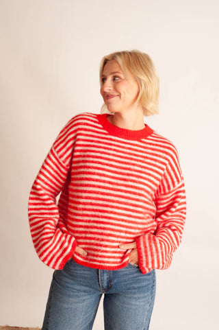 A woman with blonde hair wearing a red and white striped fuzzy knit sweater and blue jeans, standing against a plain off-white background.