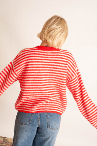 A woman with short blonde hair is shown from behind wearing a red and white striped knit sweater and blue jeans against a plain off-white background.