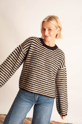 A woman wearing a fuzzy, long-sleeve knit sweater with horizontal black and beige stripes, paired with blue jeans, posing against a plain off-white background.