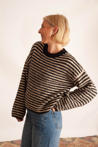 A woman with blonde hair wearing a black and beige striped fuzzy knit sweater and blue jeans, standing against a plain off-white wall.