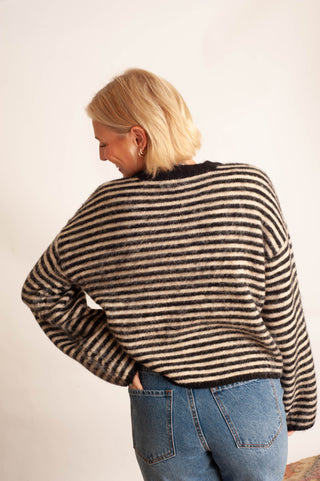 A woman with blonde hair is shown from behind wearing a black and beige striped knit sweater and blue jeans against a plain off-white background.