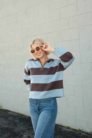 A woman wearing a light blue and dark brown striped half-zip pullover sweater with blue jeans, standing against a light gray brick wall.