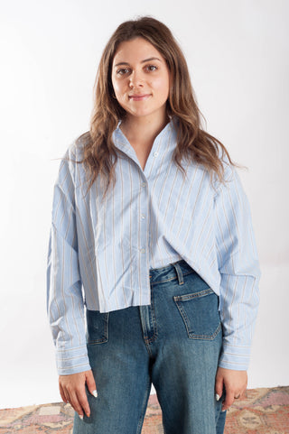 A woman modeling a light blue and white vertically striped button-up shirt with a cropped, asymmetrical hem, paired with blue jeans, against a plain white studio background.