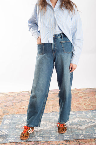 A person wearing loose-fit medium-wash blue jeans, a light blue and white striped button-up shirt, and leopard-print slip-on shoes with orange laces, standing on a patterned rug against a white studio background.