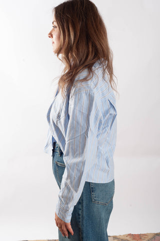A woman shown in profile wearing a light blue and white vertically striped button-up shirt with blue jeans, photographed against a plain white studio background.