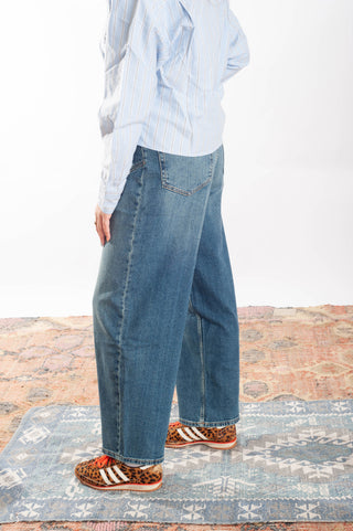 A person wearing wide-leg blue denim jeans, a light blue and white striped button-up shirt, and leopard-print sneakers with white stripes, standing on a patterned rug against a white wall.