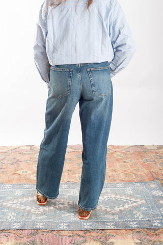 A person shown from the back wearing wide-leg blue jeans, a light blue and white striped button-up shirt, and brown shoes, standing on a patterned rug against a white wall.