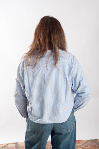 A person viewed from behind wearing a light blue and white vertically striped button-up shirt and blue jeans, standing against a plain white wall.