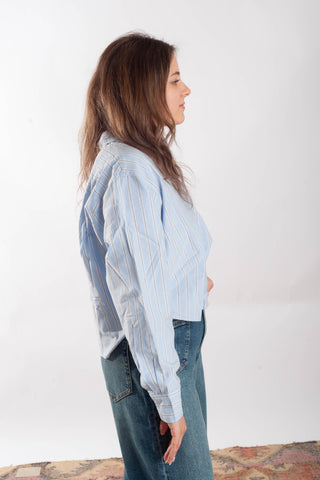 A woman in profile wearing a light blue and white vertically striped button-up shirt and blue jeans, standing against a plain white studio background.