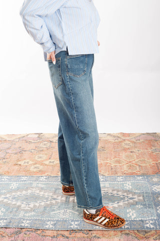 A person shown from the waist down wearing blue straight-leg denim jeans, a light blue and white striped long-sleeve shirt, and leopard-print sneakers with white stripes, standing on a patterned rug against a white wall.