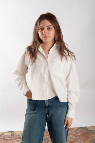 A woman wearing a white long-sleeve button-up shirt and blue jeans, standing against a plain white studio background.