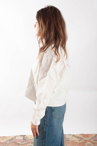 A woman shown from the side profile wearing a loose-fitting, off-white long-sleeve blouse and blue jeans, standing against a plain white studio background.