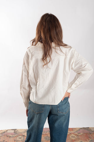A woman viewed from behind wearing a loose, long-sleeve off-white shirt and blue jeans, standing against a plain white studio background.