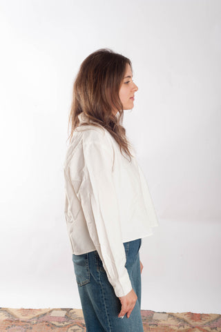 A woman in profile wearing a loose, off-white long-sleeve blouse and blue jeans, standing against a plain white studio background.