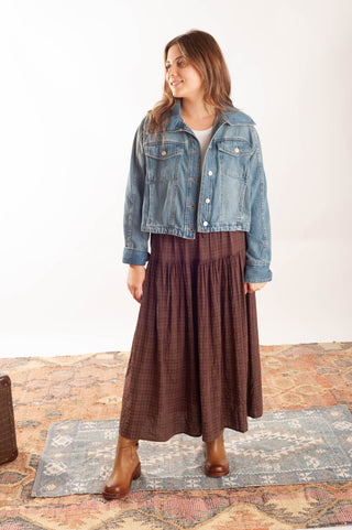 A woman modeling a cropped light-wash blue denim jacket over a white top and a brown plaid maxi skirt, standing on a patterned rug against a white studio background.